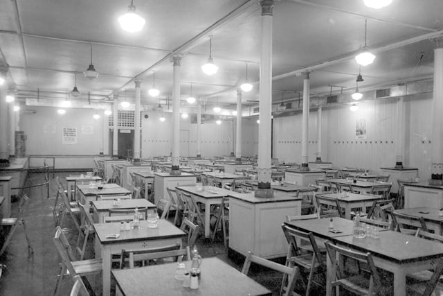 The dining room of a British Restaurant, a cheap public eating place during World War II, at Leeds Town Hall in August 1942. Rows of tables are set out with salt and pepper and ashtrays. Signs ask for plates to be returned and an advertisement for markets is visible on a wall on the right.