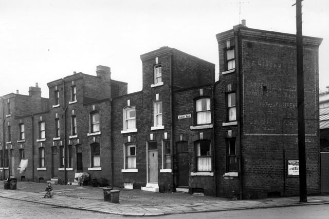 This view looks from Clarence Road onto unusual properties on Albury Road. Each house has two levels, one of three and one of two storeys. Doors to the side of the chimneys on the three storey sections give access to the rooves of the two storey sections. Houses have unfenced yards to the front where several dustbins are visible along with a small boy on a tricycle. Pictured in October 1958.