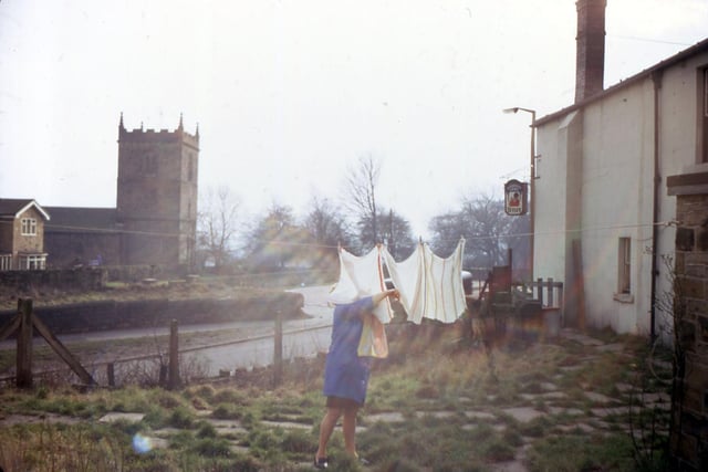 A woman is pegging washing on a line in . Behind her is Dewsbury Road and Woodkirk Church (St. Mary's) in February 1971.