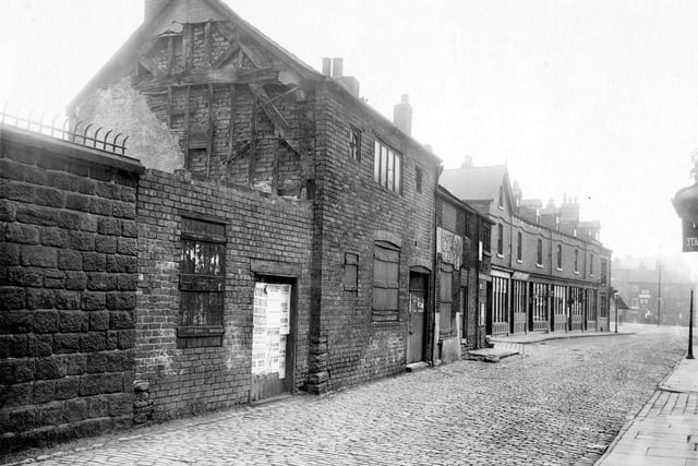 Mabgate showing the high boundary wall of St. Mary's Church on the left, followed by a row of derelict buildings, very old cruck framed timber houses which could date from the medieval period. The entrance to St Mary's Street is seen next, then a row of shops, the nearest one being Isaac and Grace Rhudstein, cap makers, at number 28 Mabgate. At the end of the row of shops is Quarry Hill (Tunstalls Fold). On the right the City of Mabgate Inn is just visible. Pictured in September 1919.