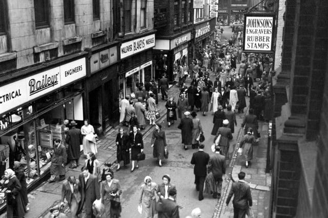 An elevated view looking south down Trinity Street in Julky 1956. On the left are N.G. Bailey & Co. Ltd., electrical engineers;  Blakeys Wallpapers; then George And Dragon Yard; J. Graham (Leeds) Ltd., home furnishers and Baker's Household Stores. On the right a sign for Johnson's Engravers (Leeds) Ltd (number 12) can be seen.