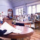 Inside Armley Branch Library on Stocks Hill in June 1985. In the foreground is a reading table where a man is sitting reading a newspaper. Behind this is a display table with a stand of records and cassettes on the right. In the background is the counter area and entrance.