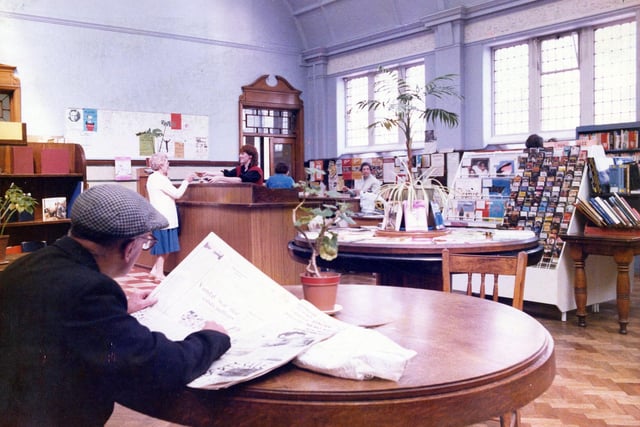 Inside Armley Branch Library on Stocks Hill in June 1985. In the foreground is a reading table where a man is sitting reading a newspaper. Behind this is a display table with a stand of records and cassettes on the right. In the background is the counter area and entrance.