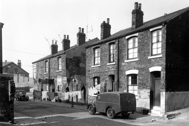 A row of four back-to-back terraced houses on Eastwood Street separated between numbers 3 and 5 by a shared outside toilet block. Clothes hang on lines stretched across the street. At the far end of the street is Cross Stamford Street where an advertisement for Senior Service cigarettes is visible. This area was known locally as Newtown. Pictured in September 1959.