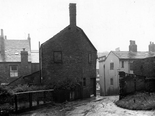 The Gang located at the back of Town Street. A muddy road is visible leading up to to the foreground from Town Street. The backs of houses are visible on the left with a garden and bushes etc. The premises of 'Isaac Stephenson Ltd. Butchers' can be seen through the gap. A streetlamp is visible to the right of the photo. Pictured in August 1952.