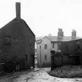 The Gang located at the back of Town Street. A muddy road is visible leading up to to the foreground from Town Street. The backs of houses are visible on the left with a garden and bushes etc. The premises of 'Isaac Stephenson Ltd. Butchers' can be seen through the gap. A streetlamp is visible to the right of the photo. Pictured in August 1952.
