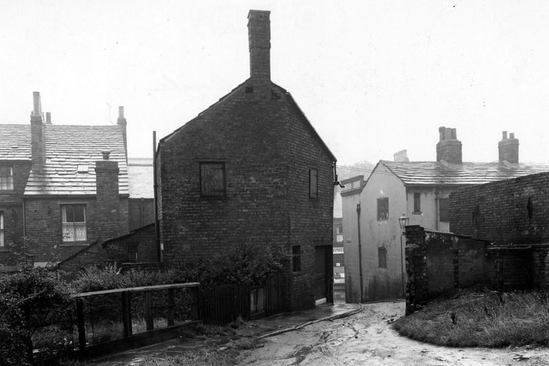 The Gang located at the back of Town Street. A muddy road is visible leading up to to the foreground from Town Street. The backs of houses are visible on the left with a garden and bushes etc. The premises of 'Isaac Stephenson Ltd. Butchers' can be seen through the gap. A streetlamp is visible to the right of the photo. Pictured in August 1952.