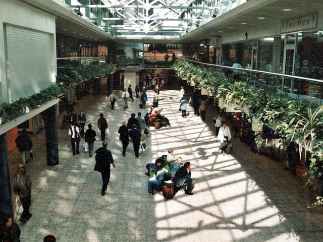 Morrisons is partially visible in the background and Inches on the right. The area is full of shoppers with benches and litter bins down the centre of the aisle.