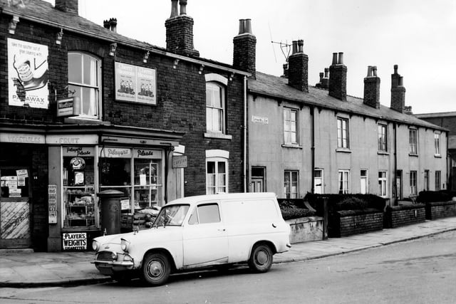 A grocers on Cotton Mill Row pictured in February 1966. The shop was closed on Tuesdays according to a sign in the door. Outside next to a post box marked 'Postal District Number 10' crates and sacks are stacked while a van is parked on the street. On the right is a row of through terraced houses with private front gardens.