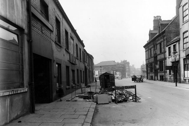 Roadworks on Hunslet Road leading up to the junction with South Brook Street. A workman stands in a hut by "road closed" signs and a cordoned off hole. Moat Engineering, Doyle and Co. Ltd. financial agents and Roy (roofing) Ltd. are visible. Two people getting out of a car and a row of advertisements are in the background. Street lamps line the street.
