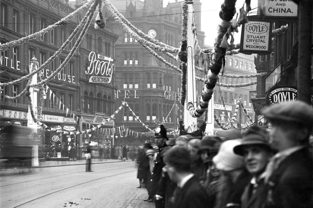 This view looks across a garland-strewn Boar Lane towards Boots the Chemists and the Griffin Hotel. Pedestrians line the street while overhead are streamers and bunting with a crown as the centrepiece.