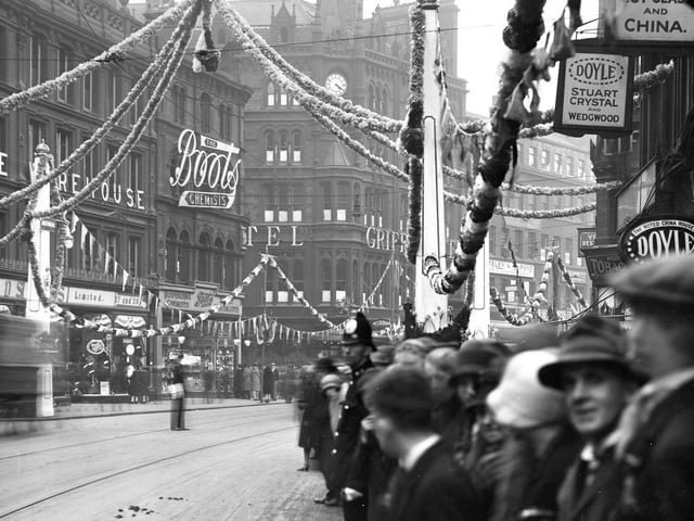 This view looks across a garland-strewn Boar Lane towards Boots the Chemists and the Griffin Hotel. Pedestrians line the street while overhead are streamers and bunting with a crown as the centrepiece.