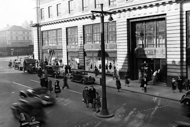 The north east side of The Headrow, showing Lewis's Department Store at numbers 22 to 26. The elevated view shows cars and a van parked and shoppers crossing the road. A man is up a ladder on the left. Some graffiti can be seen on Wade Lane. Traffic lights and a double lampost are prominent. Pictured in March 1949