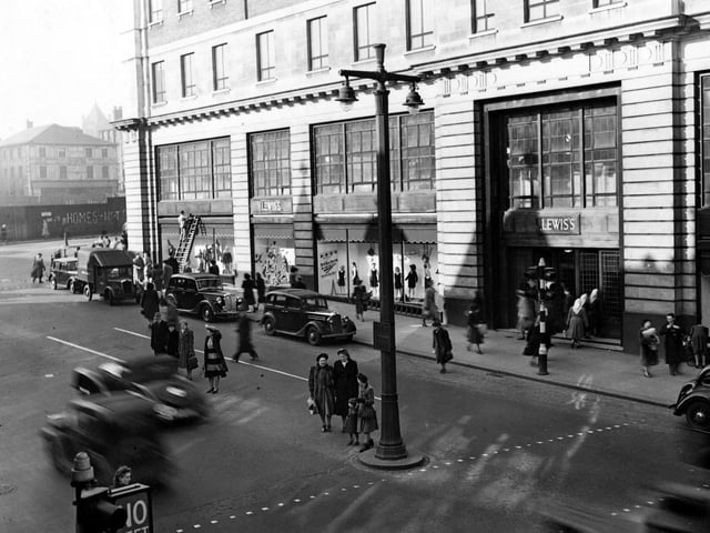 The north east side of The Headrow, showing Lewis's Department Store at numbers 22 to 26. The elevated view shows cars and a van parked and shoppers crossing the road. A man is up a ladder on the left. Some graffiti can be seen on Wade Lane. Traffic lights and a double lampost are prominent. Pictured in March 1949