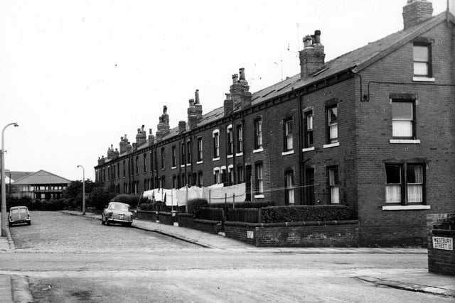 Washing lines hang across the small front gardens on Westbury Street. At the end of the road, part of a large warehouse or industrial property can be seen.