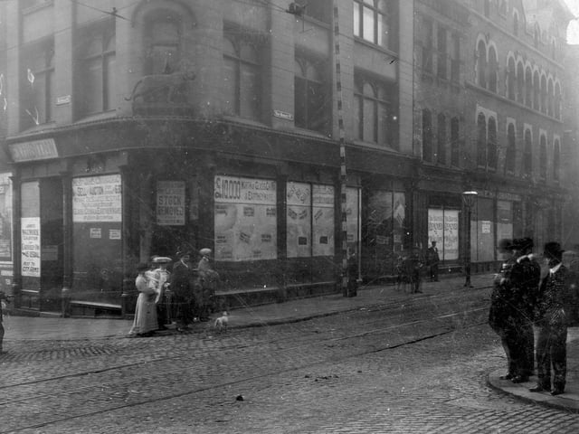The junction of New Market Street with Kirkgate former premises of Tiger Stores. Statue of tiger sited above doorway on corner. Row of empty business premises. Women, men and children in period dress pose on corners of roadside. Pictured in July 1907.