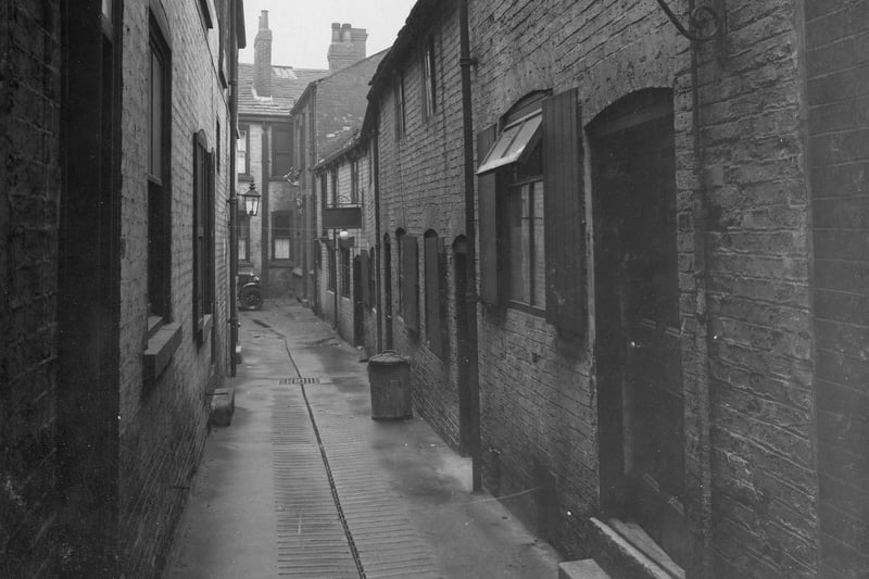 This view is down the length of Bramleys Yard off The Headrow in June 1936 was described as an unhealthy area on the photograph notes. At the bottom and on the right side the Arm and Hammer emblem of the Gold Beaters Guild shows where the shop of W.Barnes is situated. The yard also contains the staff and goods entrance of J. Lyons &Co.