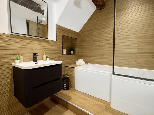 The family bathroom features a P-shaped bath, which is set upon a plinth and fitted with an overhead shower. Black fixtures add a sense of style and sophistication, with a two-drawer vanity unit wash basin and WC.