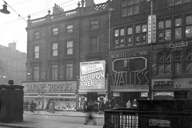 Briggate with the junction with Boar Lane on the left. In the foreground is a police telephone box and the entrance to the gentlemen's public toilets. The shops shown are; Number 33, Saxone & Sorosis Shoe Co. Ltd.; Number 35 is Edmond's wool shop, who are having a 'coupon free stock clearance'. Number 36 is Wallis & Co (Costumiers) Ltd, (who are having a winter sale). Number 37 is S. Tetley & Sons Ltd, tobacconist, who have a window advertisement for Gold Flake. Above number 37 is Franks (Opticians) Ltd. Pedestrians are visible on the pavement.
