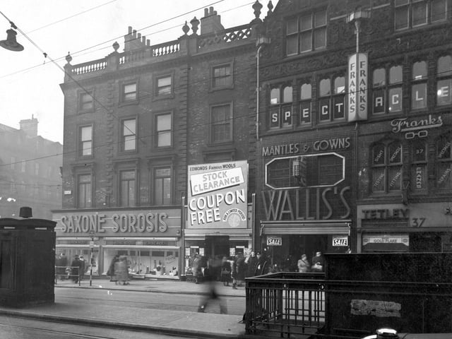 Briggate with the junction with Boar Lane on the left. In the foreground is a police telephone box and the entrance to the gentlemen's public toilets. The shops shown are; Number 33, Saxone & Sorosis Shoe Co. Ltd.; Number 35 is Edmond's wool shop, who are having a 'coupon free stock clearance'. Number 36 is Wallis & Co (Costumiers) Ltd, (who are having a winter sale). Number 37 is S. Tetley & Sons Ltd, tobacconist, who have a window advertisement for Gold Flake. Above number 37 is Franks (Opticians) Ltd. Pedestrians are visible on the pavement.