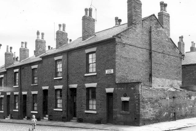 A small boy rides his scooter on Acton Street from the junction with Kiln Street. The end door and window on the right are those of a block of several outside toilet facilities for the use of the street. It was an area designated for clearance in line with the Housing Act of 1957.