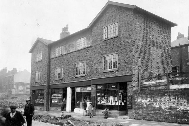 Burley Street after improvement in April 1930. Row of four shops, with accommodation over. Number 22, Mrs P. Benn, greengrocer number 20, R. Chambers, boot and shoe repairs. Number 18 J. Turnbull, butcher and number 16 Alice Cordingley, sweets and groceries. On the left, cleared area was Rutland Mount. Road is still to be finished, Tram Lines can be seen in cobbled portion. There is a vending machine for nuts and raisins on the wall outside sweet shop, children in view.
