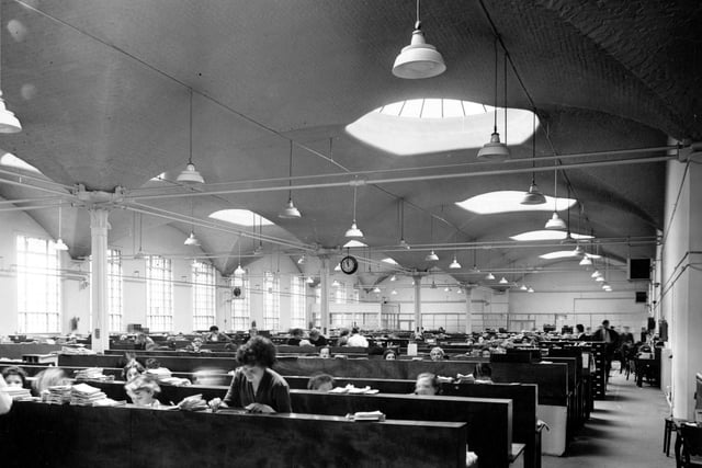 Inside Marshall's Mills in August 1956. Rows of desks are in a large office with support posts running down the middle. People sit at the desks and papers are piled up. A clock hangs from the ceiling. Windows and skylights are visible.