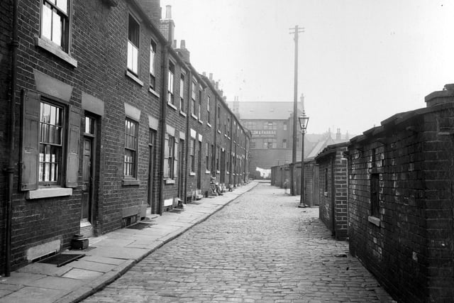 A row of terraced houses on Lilac Terrace in August 1935 with outside privies opposite. Windows have wooden shutters. Women sitting on front step further down. Gas lamp in street, which is paved with stone sets. At the end of the road can be seen Walton and Farrar, manufacturing upholsterers, on Concord Street.