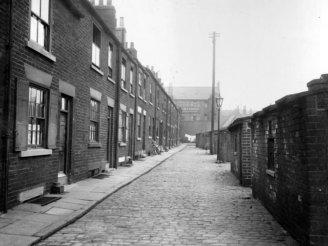 A row of terraced houses on Lilac Terrace in August 1935 with outside privies opposite. Windows have wooden shutters. Women sitting on front step further down. Gas lamp in street, which is paved with stone sets. At the end of the road can be seen Walton and Farrar, manufacturing upholsterers, on Concord Street.