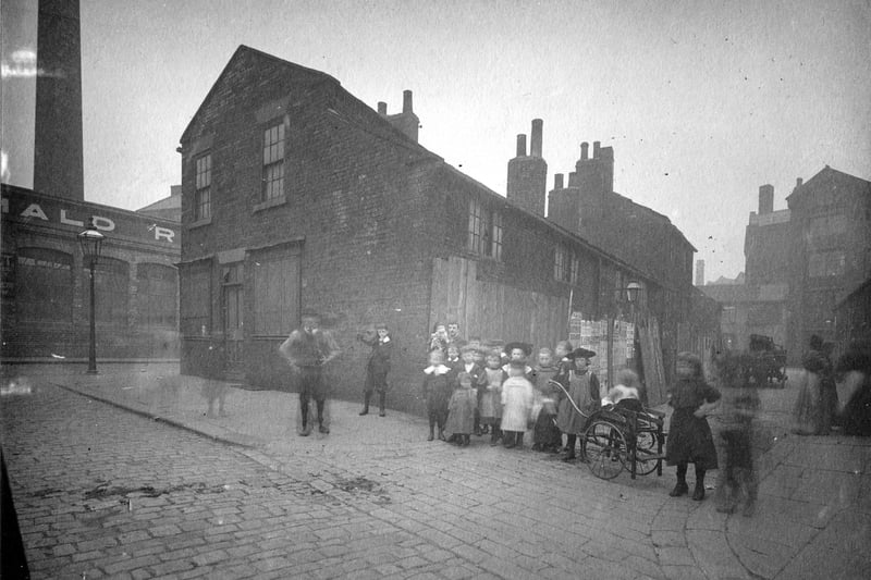 Property clearance for improvements to Park Lane, the buildings to be demolished are behind fencing. A group of children in period dress are posing for the camera, one has a push cart.