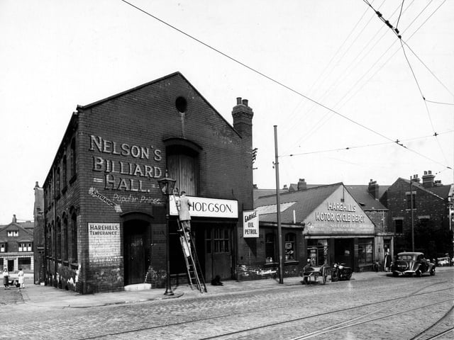 'Nelsons Billiard Hall' and 'Harehills Motorcycle Depot', with 'Hodgsons' in between on Harehills Road. A man is stood on a ladder. Signs for 'Garage,Oil and Petrol', 'Plumbing', 'Spot Cash Paid', 'Harehills Temperance Club' among others visible. Car, motorcycle, cart visible to the right. Two ladies to the left of the picture with the 'Provincial Bank' behind them. Pictured in August 1947.