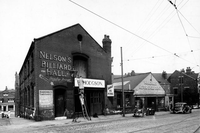 'Nelsons Billiard Hall' and 'Harehills Motorcycle Depot', with 'Hodgsons' in between on Harehills Road. A man is stood on a ladder. Signs for 'Garage,Oil and Petrol', 'Plumbing', 'Spot Cash Paid', 'Harehills Temperance Club' among others visible. Car, motorcycle, cart visible to the right. Two ladies to the left of the picture with the 'Provincial Bank' behind them. Pictured in August 1947.