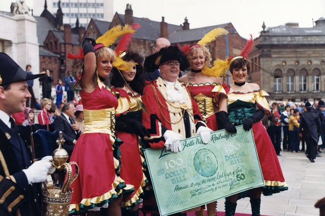 Four 'saloon girls' from Lewis's department store have left their American Wild Western 'steam engine' in the 7th Lord Mayor's Parade to present the Lord Mayor, Councillor Eric Atkinson MBE with a one hundred dollar bill. The parade has temporarily halted in front of the Civic Hall (seen left) and the Lord Mayor is in position to receive a salute from each entrant as they pass by. The Lewis's girls are attired in red dresses trimmed with green and gold, black fishnet stockings and buttoned boots. The outfits are accessorised with black, elbow length gloves, black chockers and sequinned heddresses decorated with red and yellow ostrich feather. The Mace bearer is seen at the left edge and there is a good view of the Leeds Civic Mace which dates back to 1694, and was made by Arthur Mangey, a goldsmith of Briggate.