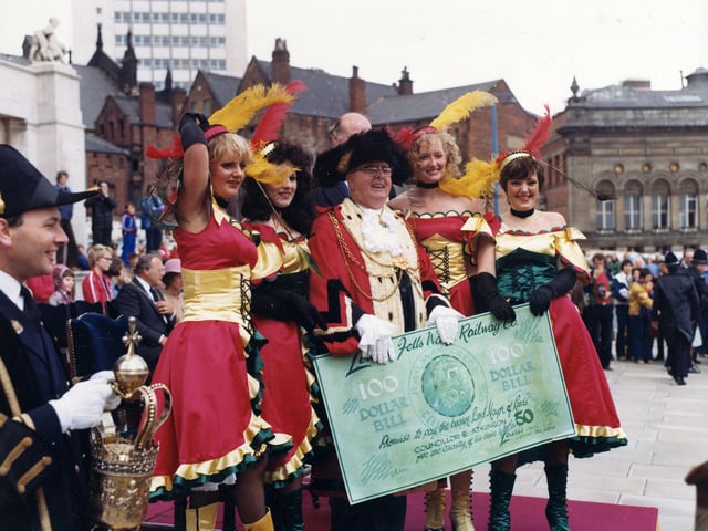 Four 'saloon girls' from Lewis's department store have left their American Wild Western 'steam engine' in the 7th Lord Mayor's Parade to present the Lord Mayor, Councillor Eric Atkinson MBE with a one hundred dollar bill. The parade has temporarily halted in front of the Civic Hall (seen left) and the Lord Mayor is in position to receive a salute from each entrant as they pass by. The Lewis's girls are attired in red dresses trimmed with green and gold, black fishnet stockings and buttoned boots. The outfits are accessorised with black, elbow length gloves, black chockers and sequinned heddresses decorated with red and yellow ostrich feather. The Mace bearer is seen at the left edge and there is a good view of the Leeds Civic Mace which dates back to 1694, and was made by Arthur Mangey, a goldsmith of Briggate.