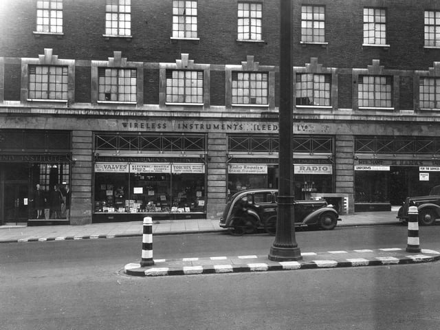 Leeds Workshops for the blind shop selling goods made by the visually impaired. Number 54/56 Wireless Instruments Leeds Ltd, selling radios, torches, batteries, valves for radios and parts, repairs and servicing. Number 52, H. Banks, tailors dealing in uniforms for servicemen and women. Notices in window state 'Uniforms in all sizes', 'Tropical Kit in stock'. White lines on road, central road island and bollards acted as visual aid during war-time black out. Pictured in April 1942.