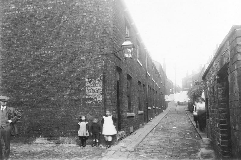 Looking north along Back Portland Crescent near the south entrance to St. James' Square in September 1913. Grates can be seen over cellar windows on the left of the narrow, cobbled street. A man and other blurred figures are visible. Yards with outside toilets are on the right. Washing hangs up and streetlamps can be seen. The area is now the site for Civic Hall.