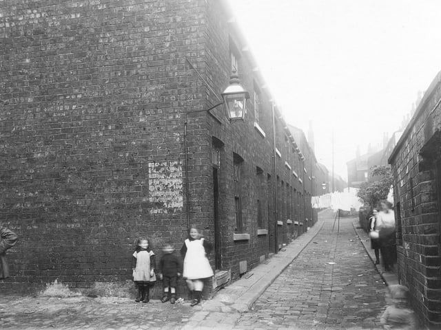 Looking north along Back Portland Crescent near the south entrance to St. James' Square in September 1913. Grates can be seen over cellar windows on the left of the narrow, cobbled street. A man and other blurred figures are visible. Yards with outside toilets are on the right. Washing hangs up and streetlamps can be seen. The area is now the site for Civic Hall.