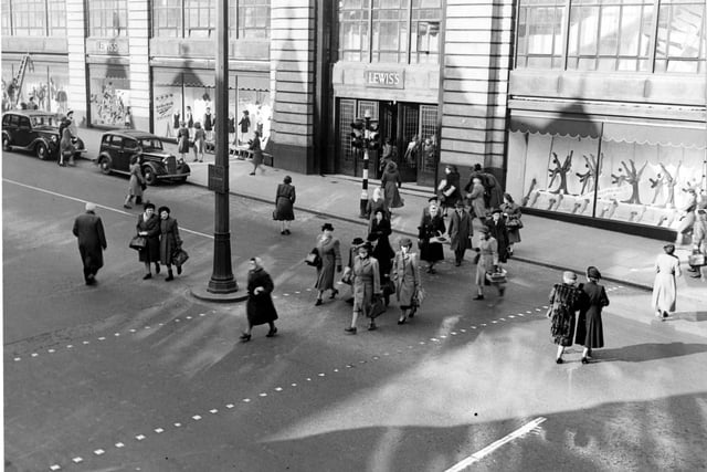 Pedestrians cross the road outside Lewis's (Leeds) Ltd. department store in March 1949. A ladder is leant against the store and cars are parked outside.