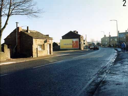 Bradford Road in December 1990, showing a fish and chip shop at no. 28 on the left. Next to this is an unmade parking area, then an advertising hoarding for Tetley's Bitter can be seen on the side of no. 22. In the distance on the right is the Victoria public house, at the junction with Whitehall Road.