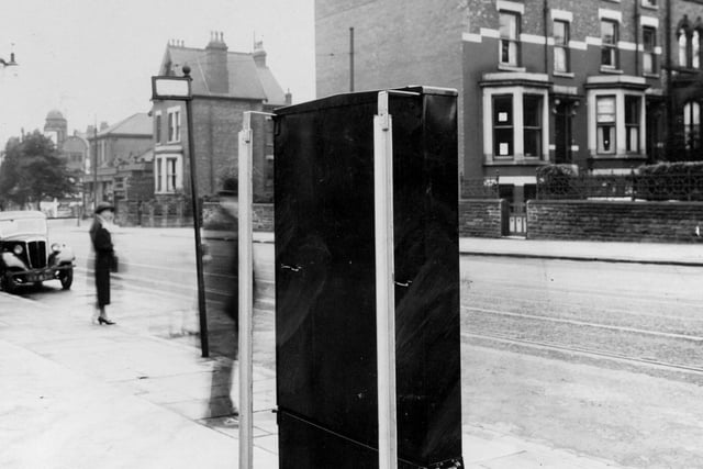 A cigarette machine on Chapeltown Road pictured in August 1939. To the left of the photo is a parked car, and a woman standing on the pavement. Near the cigarette machine is a tram stop.