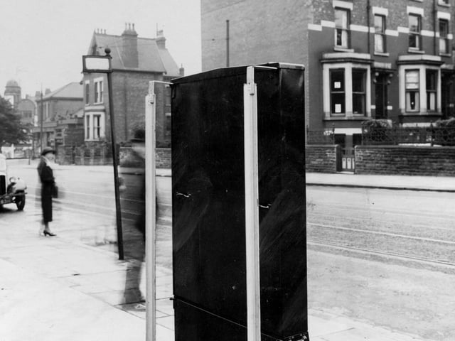 A cigarette machine on Chapeltown Road pictured in August 1939. To the left of the photo is a parked car, and a woman standing on the pavement. Near the cigarette machine is a tram stop.