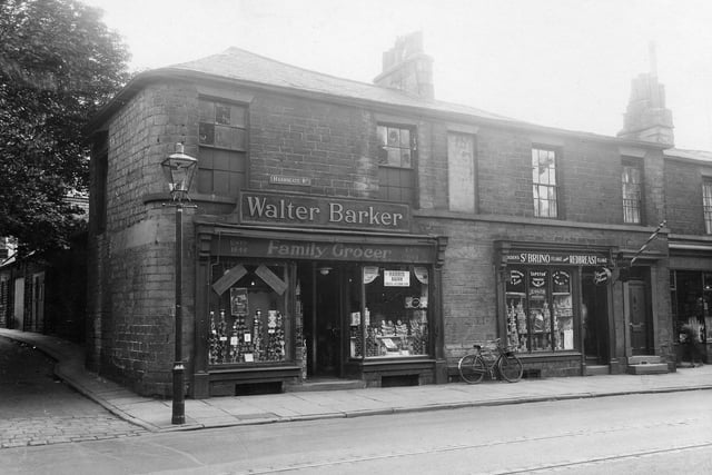 Harrogate Road at the junction with Short Lane in August 1935. Here is a row of stone built shops with slate roofs. On the corner is W.Barker, Family grocers, Est 1844. Next is J.E.Bolton, Hairdresser. In the window of this shop there are advertisments for St.Bruno and Redbreast Flake, pipe tobaccos. A gas streetlamp stands on the pavement and a trademans bike leans against the wall.