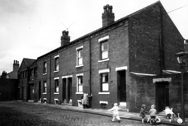 A row of back-to-back terraced houses on Temple View Terrace with warehouse buildings on the left and a shared outside toilet block on the right. A man and a woman are stood outside number 87 while children play on tricycles on the right. Pictured in July 1963.