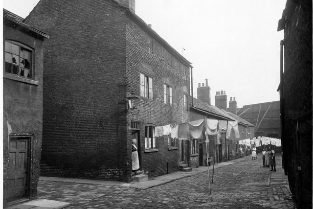 The entrance to Chadwick Court and residential accommodation on one side of the street. There are three lines of washing hanging over the street and several people watching the photographer. In the foreground there is a dilapidated building on the left next to the entrance to the court. Pictured in October 1935.