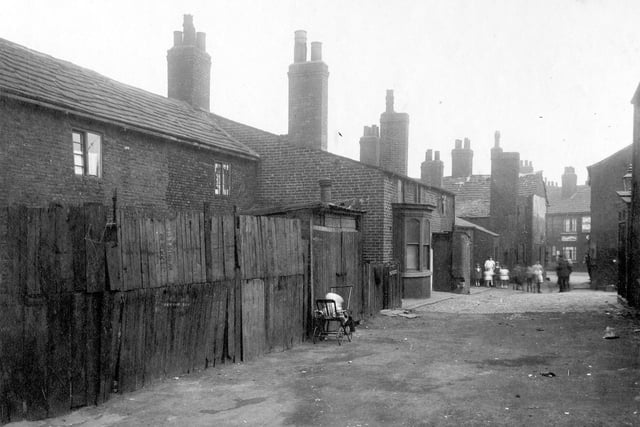 Finny Yard was a conglomeration of properties. To the left, fencing and shed, child in push chair. A group of children stand near the yard entrance.