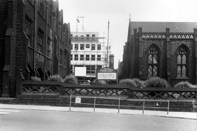 Looking east in July 1956 towards Mill Hill Unitarian Chapel, the arched leaded windows of which are visible. In the centre is the chapel's "wayside pulpit" a notice with a quote on it. On the left are the large stone premises of The Ocean, Accident & Guarantee Corp. Ltd, at number 1. In the background can be seen a new building under construction. In front of the chapel's ornate stone wall is a pedestrian barrier.