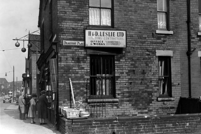 A view of Roundhay Road at the junction with Beaufort Place on the south east side of the road ion September 1953. The end of Badminton Street is visible. A group of people look in the window of Harry Waite's pawnbrokers. A sign directs towards H. & D. Leslie building contractors. A tram and a car are on the road.