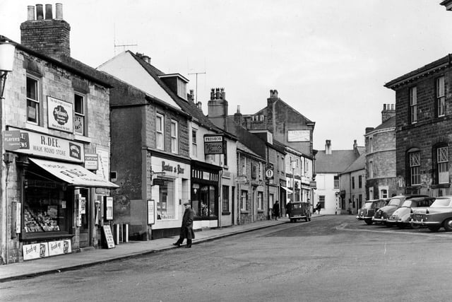 The Market Place looking east towards the High Street in June 1962. Pictured, far left,  at number 45 Market Place, is R. Dee's 'Walk Round Store', Taxis and Newsagent. Various advertisements are displayed, including one for Neilson's Canadian Icecream. Adjacent to Dee's is an alleyway which cuts through to Bank Street. Moving right is estate agents, Renton & Parr at number 47, then Preston's Wines and Beers,. The Black Bull is at number 53, and, further along the Gas showrooms. The Town Hall is located at the right edge.