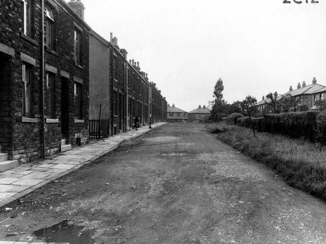 Looking north along Nora Place towards the junction with Leeds and Bradford Road in August 1954. The road is unsurfaced with a grassy area and hedge covered fence down the middle. An old woman and streetlamps are visible.