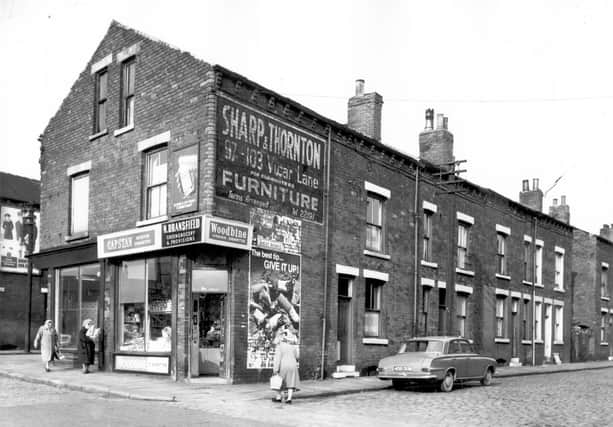 A shop at junction of Ascot Avenue and Pontefract Lane. Two women walk towards the N. Dransfield greengrocery & provisions store from the left, while another woman crosses the street on the right to reach the store. Above the shop are two advertisements for Dettol on the left and Sharp & Thornton furniture store on the right. On the right a row of back-to-back terraces are visible with a car, reg: 408 DUM parked outside. Pictured in October 1966.